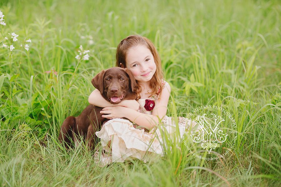 Portrait of a little girl with her chocolate lab puppy in a pretty green field in St. Louis taken by Yvonne Niemann Photography.