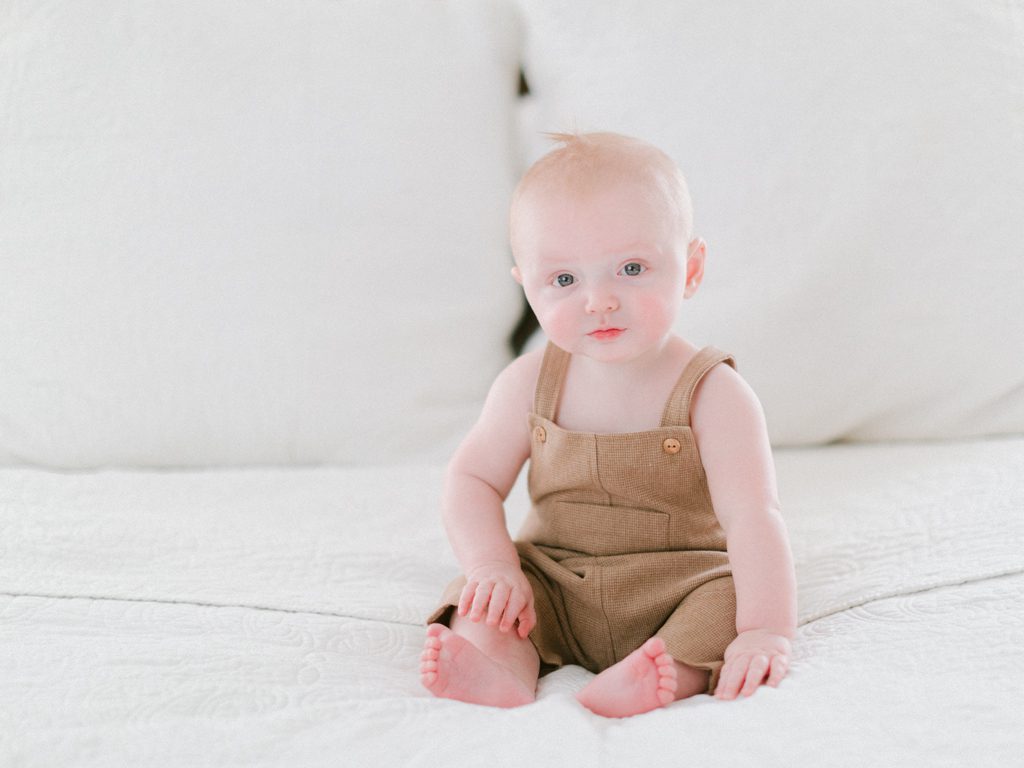 Photo of a seven month old baby sitting on a bed with white linens wearing a Zara romper. Taken in Oconee County, GA.